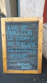 A wooden-framed blackboard propped up on a sidewalk displays a quote in light blue chalk. It is positioned against a beige and red wall.