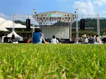 An outdoor event features a stage set up with large speakers on each side, surrounded by a grassy area. Several people are sitting on the grass facing the stage. The backdrop behind the stage is clear, and the sky is mostly blue with a few clouds. Trees and hills are visible in the distance, adding a natural setting to the scene.