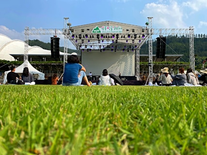 An outdoor event features a stage set up with large speakers on each side, surrounded by a grassy area. Several people are sitting on the grass facing the stage. The backdrop behind the stage is clear, and the sky is mostly blue with a few clouds. Trees and hills are visible in the distance, adding a natural setting to the scene.
