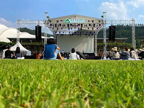 An outdoor event features a stage set up with large speakers on each side, surrounded by a grassy area. Several people are sitting on the grass facing the stage. The backdrop behind the stage is clear, and the sky is mostly blue with a few clouds. Trees and hills are visible in the distance, adding a natural setting to the scene.
