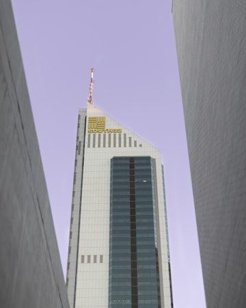 A tall, modern skyscraper with a prominent glass facade and a sharp, angular design. The building is positioned between two other tall structures and features the logo 'SOUTH32' near its top. The sky is a pale, muted color, providing a stark contrast to the building's sleek surfaces.