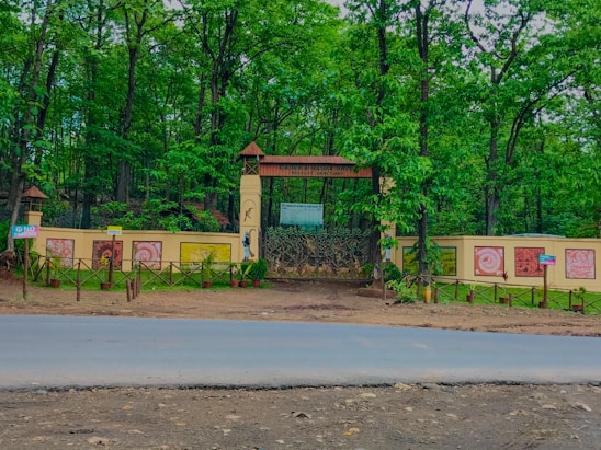 A gated entrance surrounded by lush green trees, with elaborate designs and murals on the walls. The pathway is bordered by a small wooden fence and potted plants. A paved road runs in front of the entrance.