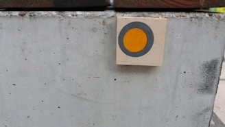 A light-colored concrete block with small speckles and visible texture is in the foreground. A small wooden square with a painted circle, featuring a gray outer ring and an orange center, is mounted on the right side of the block.
