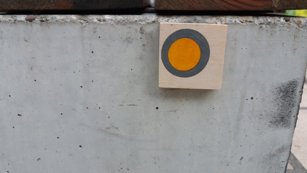 A light-colored concrete block with small speckles and visible texture is in the foreground. A small wooden square with a painted circle, featuring a gray outer ring and an orange center, is mounted on the right side of the block.