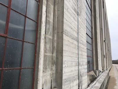 A long industrial building constructed with concrete and large grid-patterned windows. The windows are framed in red, contrasting with the gray concrete surface. The perspective emphasizes the building's length as it extends into the distance.