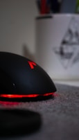 A sleek black computer mouse with red lighting accents, sitting on a gray surface. In the blurred background, there is a white container holding colorful pens or pencils, partially visible.
