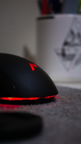 A sleek black computer mouse with red lighting accents, sitting on a gray surface. In the blurred background, there is a white container holding colorful pens or pencils, partially visible.