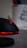 A sleek black computer mouse with red lighting accents, sitting on a gray surface. In the blurred background, there is a white container holding colorful pens or pencils, partially visible.