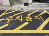 Yellow paint marks the words in Korean characters on a dark asphalt surface, framed by diagonal lines indicating a restricted parking area. The background features part of a building, a tire, and some urban elements.