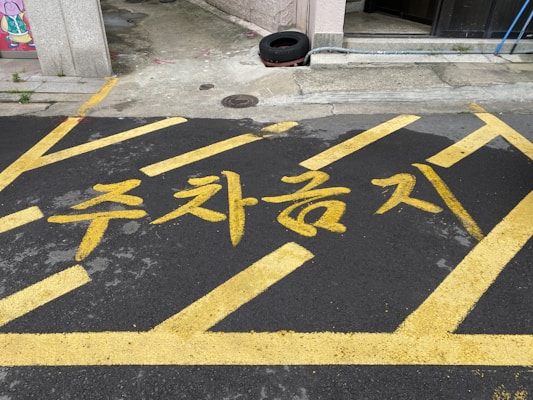 Yellow paint marks the words in Korean characters on a dark asphalt surface, framed by diagonal lines indicating a restricted parking area. The background features part of a building, a tire, and some urban elements.
