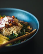 A close-up view of a bowl filled with quinoa, roasted chickpeas, chopped red onions, and leafy greens. The bowl is a deep blue color and a wooden spoon is resting inside. The ingredients are arranged with care, creating an appetizing look.