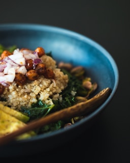 A close-up view of a bowl filled with quinoa, roasted chickpeas, chopped red onions, and leafy greens. The bowl is a deep blue color and a wooden spoon is resting inside. The ingredients are arranged with care, creating an appetizing look.