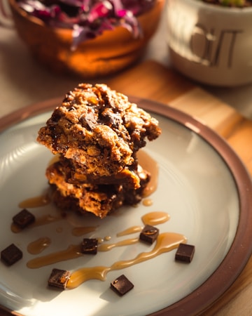 A plate holds a stack of three chocolate chip cookies drizzled with caramel sauce. Small chocolate cubes are scattered around the cookies, adding a decorative touch. The setting includes warm lighting that highlights the texture of the cookies, creating an inviting atmosphere. In the background, blurred elements such as a plant and a bowl enhance the composition.