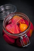 A glass jar filled with vibrant pickled vegetables, including sliced beets and a small yellow vegetable, immersed in a deep pink liquid. The jar is sealed with a metal clasp and sits on a dark surface.