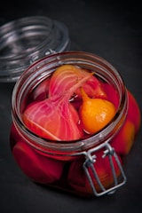 A glass jar filled with vibrant pickled vegetables, including sliced beets and a small yellow vegetable, immersed in a deep pink liquid. The jar is sealed with a metal clasp and sits on a dark surface.