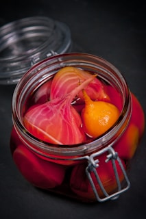 A glass jar filled with vibrant pickled vegetables, including sliced beets and a small yellow vegetable, immersed in a deep pink liquid. The jar is sealed with a metal clasp and sits on a dark surface.