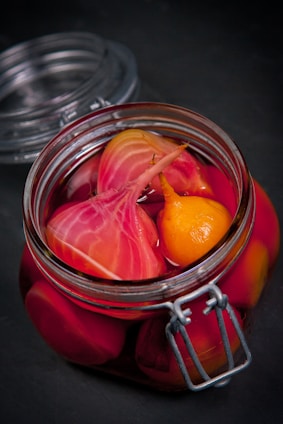 A glass jar filled with vibrant pickled vegetables, including sliced beets and a small yellow vegetable, immersed in a deep pink liquid. The jar is sealed with a metal clasp and sits on a dark surface.