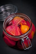 A glass jar filled with vibrant pickled vegetables, including sliced beets and a small yellow vegetable, immersed in a deep pink liquid. The jar is sealed with a metal clasp and sits on a dark surface.