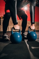 A person wearing black leggings and red shorts is holding two blue kettlebells on a gym floor with a red and black background. Light is shining from the back, casting shadows.