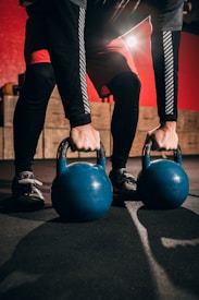 A person wearing black leggings and red shorts is holding two blue kettlebells on a gym floor with a red and black background. Light is shining from the back, casting shadows.