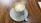 A cup of latte with heart-shaped latte art on top sits on a ceramic saucer with a spoon. The table surface is wooden, adding a warm and cozy feel to the setting.