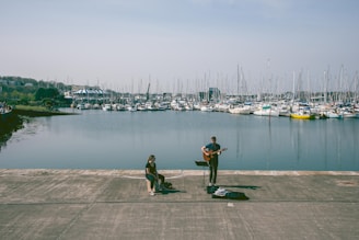 A waterfront scene featuring a musician playing a guitar and singing beside a seated listener. Numerous sailboats are docked in a marina in the background, and the water is calm under a clear sky.