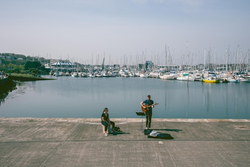 A waterfront scene featuring a musician playing a guitar and singing beside a seated listener. Numerous sailboats are docked in a marina in the background, and the water is calm under a clear sky.