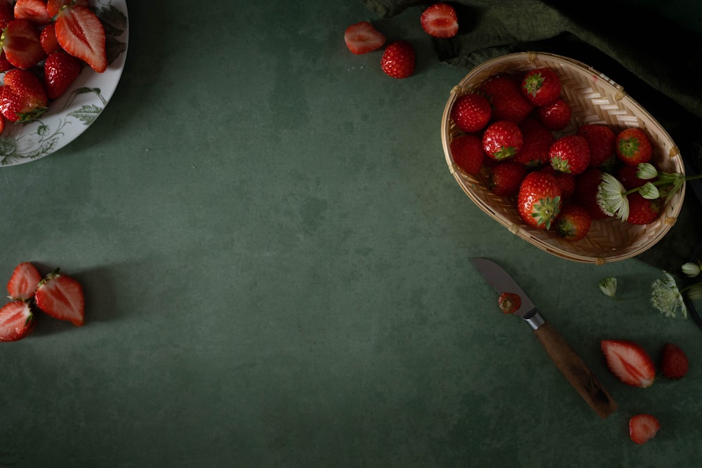 a basket of strawberries next to a knife and a bowl of strawberries