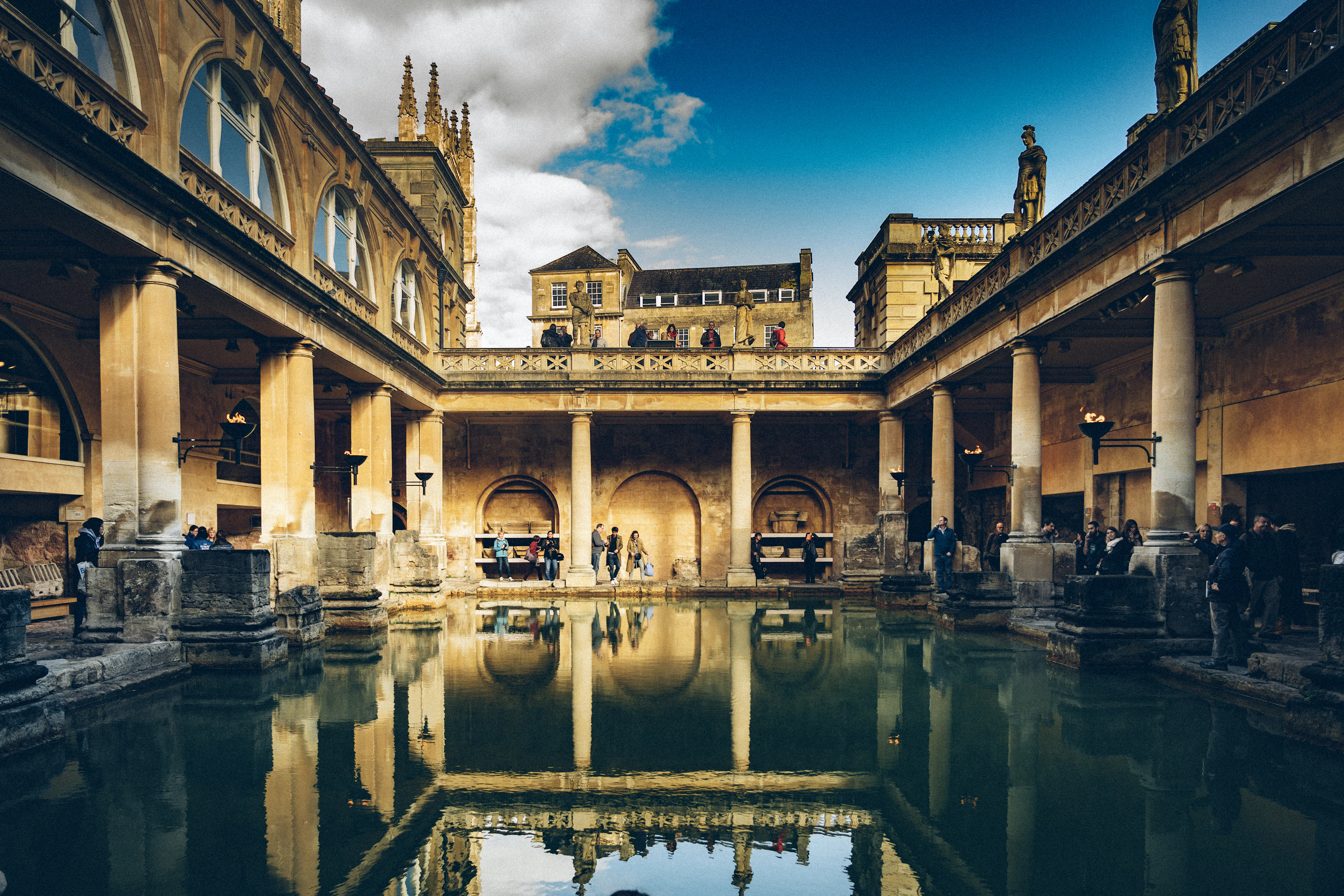 The Great Bath at the Roman Baths in Bath, its still green mineral water mirroring the honey-stone terrace of statues and the pinnacles of Bath Abbey rising behind.