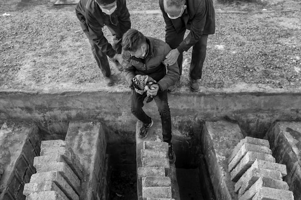 Three people are positioned near a ledge with two adults helping a younger person who appears to be in distress. The scene is set in a monochrome environment with bricks in the foreground.