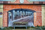 A mural painted on a brick wall creates the illusion of an arched opening leading to an old-fashioned train station interior. The painting features people walking and standing, with detailed architecture and lighting. In front, a worn bench is placed on the ground, adding depth to the scene. A clock and a no-parking sign are seen on the wall.