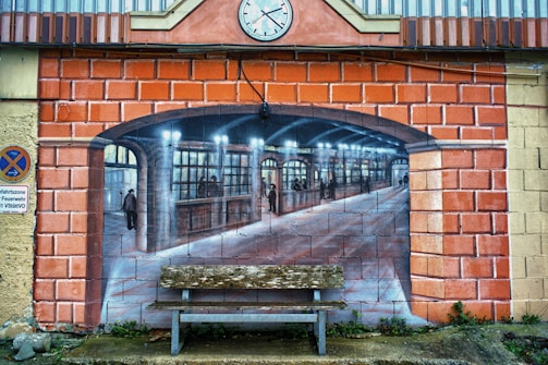 A mural painted on a brick wall creates the illusion of an arched opening leading to an old-fashioned train station interior. The painting features people walking and standing, with detailed architecture and lighting. In front, a worn bench is placed on the ground, adding depth to the scene. A clock and a no-parking sign are seen on the wall.