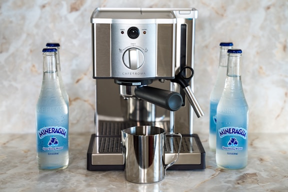 A sleek espresso machine is set on a marble countertop, surrounded by three bottles of Mineragua sparkling water. The machine, made of stainless steel, features knobs and a steam wand, with a shiny metal milk frothing pitcher placed beneath the spout. The water bottles are positioned on either side of the machine, contributing to an organized and balanced composition.