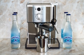 A sleek espresso machine is set on a marble countertop, surrounded by three bottles of Mineragua sparkling water. The machine, made of stainless steel, features knobs and a steam wand, with a shiny metal milk frothing pitcher placed beneath the spout. The water bottles are positioned on either side of the machine, contributing to an organized and balanced composition.