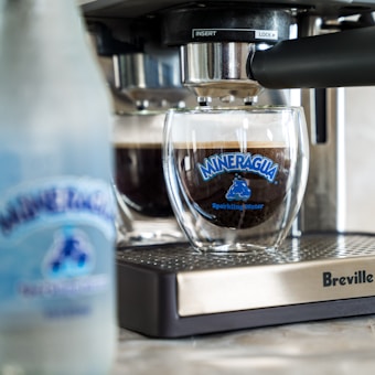 A glass cup is positioned under an espresso machine with coffee being prepared. The glass has a blue logo that reads 'Mineragua Sparkling Water.' In the foreground, there's a blurred bottle with a similar logo. The espresso machine, branded Breville, has a metallic and black finish.