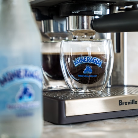 A glass cup is positioned under an espresso machine with coffee being prepared. The glass has a blue logo that reads 'Mineragua Sparkling Water.' In the foreground, there's a blurred bottle with a similar logo. The espresso machine, branded Breville, has a metallic and black finish.