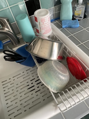 A kitchen sink with a drying rack containing various kitchen items including a stainless steel pot, a plastic container, a cup, and kitchen utensils. Nearby, there are cleaning supplies like dish soap, a sponge, and hand sanitizer on a tiled countertop.