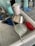 A kitchen sink with a drying rack containing various kitchen items including a stainless steel pot, a plastic container, a cup, and kitchen utensils. Nearby, there are cleaning supplies like dish soap, a sponge, and hand sanitizer on a tiled countertop.