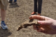 A hand holding a piece of clay or mud shaped in a sculptural form, possibly representing an animal. The background shows a dirt or mud ground with a few people wearing shorts and sneakers, slightly out of focus.