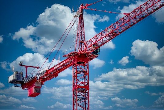 A large red construction crane extends across the sky, framed by a backdrop of white fluffy clouds. The crane has several structural parts, including a cab and a lifting boom, and is prominently labeled with branding in white. The sky is a vibrant blue, enhancing the contrast with the red crane.
