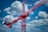 A large red construction crane extends across the sky, framed by a backdrop of white fluffy clouds. The crane has several structural parts, including a cab and a lifting boom, and is prominently labeled with branding in white. The sky is a vibrant blue, enhancing the contrast with the red crane.