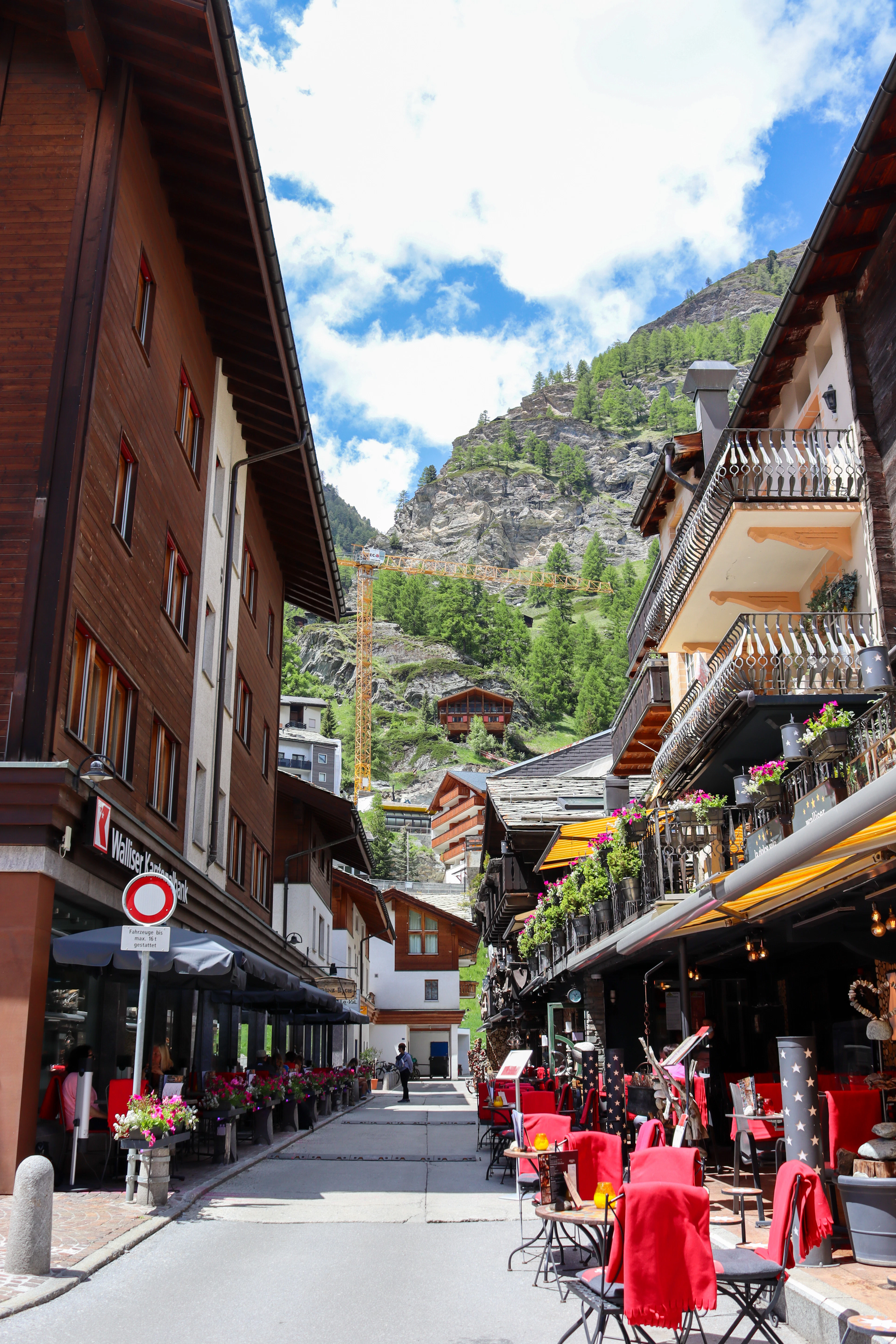 Restaurant terraces set out along a narrow street in the centre of Zermatt on a summer day, red blankets over the chairs and geraniums on the timber chalet balconies above, the pine-covered mountainside rising at the end of the lane.