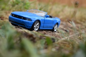 A detailed toy model of a blue convertible car sits on a dirt surface surrounded by blurred greenery. The vehicle features intricate design elements such as headlights and rims, showing a sense of realism despite being a miniature.