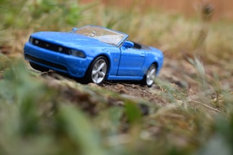 A detailed toy model of a blue convertible car sits on a dirt surface surrounded by blurred greenery. The vehicle features intricate design elements such as headlights and rims, showing a sense of realism despite being a miniature.