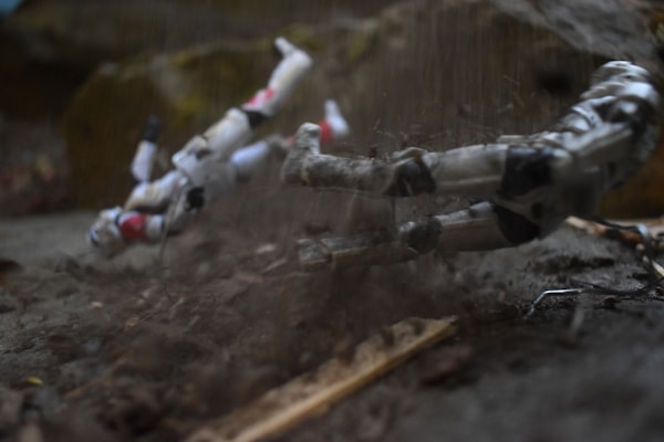 Two toy action figures resembling futuristic soldiers are depicted mid-motion, appearing to be in a dynamic action sequence. They are surrounded by loose dirt and debris, adding to the sense of movement and impact.