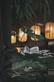 Softly glowing lanterns hang amidst lush green foliage in a dimly lit interior space. Bamboo leaves partially obscure the view, creating a natural frame around the warm amber light of the lanterns.