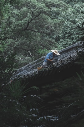 A person wearing a broad-brimmed straw hat and blue clothing is working on the tiled roof of a building. The surrounding environment is lush with dense, green foliage and tall trees, creating a serene and natural setting.