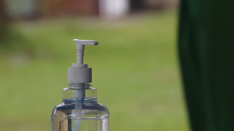 Close-up of a clear glass bottle with a natural liquid detergent surrounded by fresh green leaves on a white background