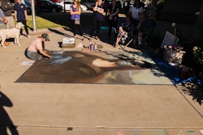 An artist is creating a large chalk drawing on the pavement, depicting a replica of the Mona Lisa. The artwork is surrounded by a small crowd of people observing. The scene is outdoors in a sunny area, with some shadows cast on the drawing. Art supplies are visible on the ground, including pastels and spray bottles.