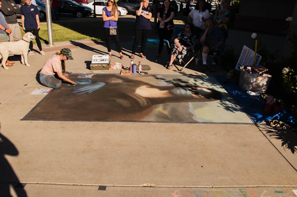 An artist is creating a large chalk drawing on the pavement, depicting a replica of the Mona Lisa. The artwork is surrounded by a small crowd of people observing. The scene is outdoors in a sunny area, with some shadows cast on the drawing. Art supplies are visible on the ground, including pastels and spray bottles.